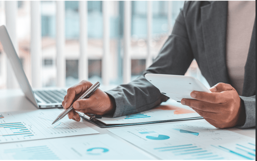 business person with a calculator in hand looking at charts next to an open lap top