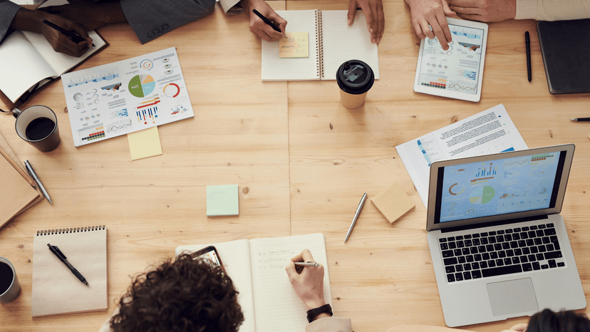 multiple people at a meeting table with notebooks, laptops, notes, and coffee, as shown from above
